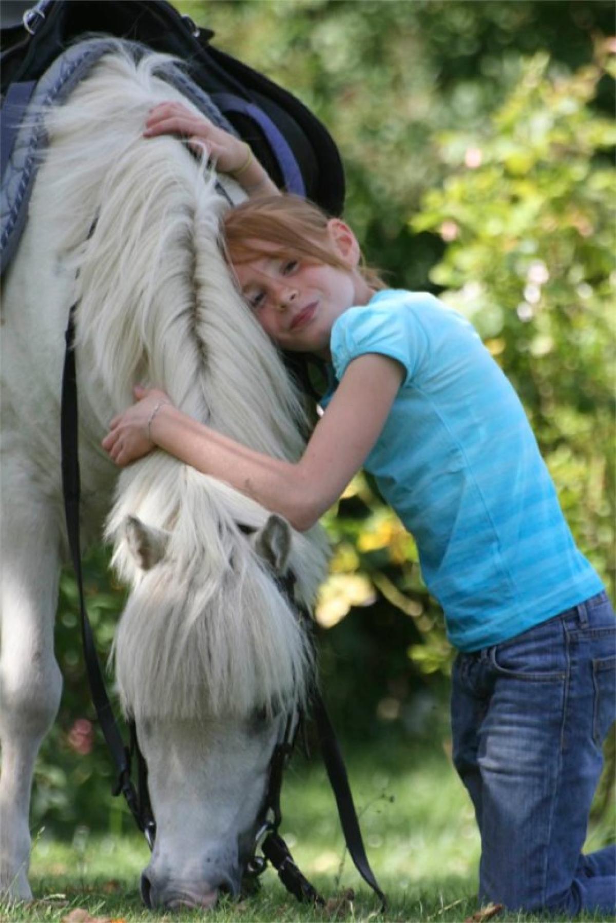 Une jeune fille rousse en t-shirt bleu enlace tendrement la crinière d'un poney blanc qui broute.